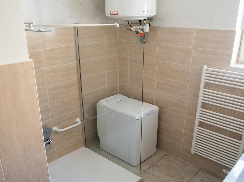 Salle de bain carrelée beige avec machine à laver à chargement par le dessus placée dans un coin douche vitré