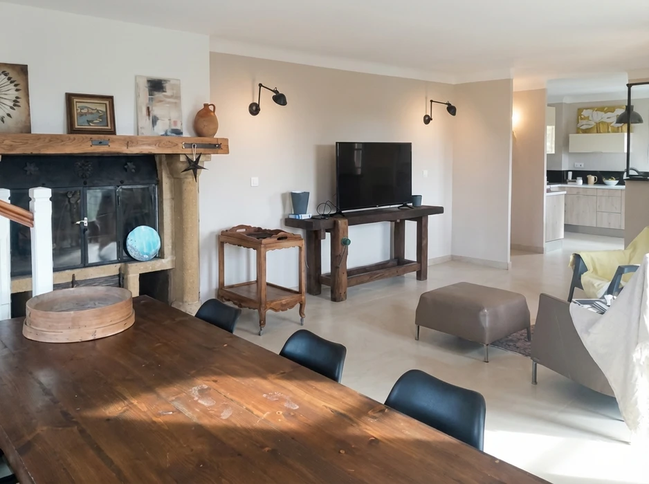 Salon ouvert avec cheminée ancienne en pierre et bois brut, table de salle à manger en bois massif, chaises noires, TV à écran plat sur console en bois rustique, ottomane en cuir taupe