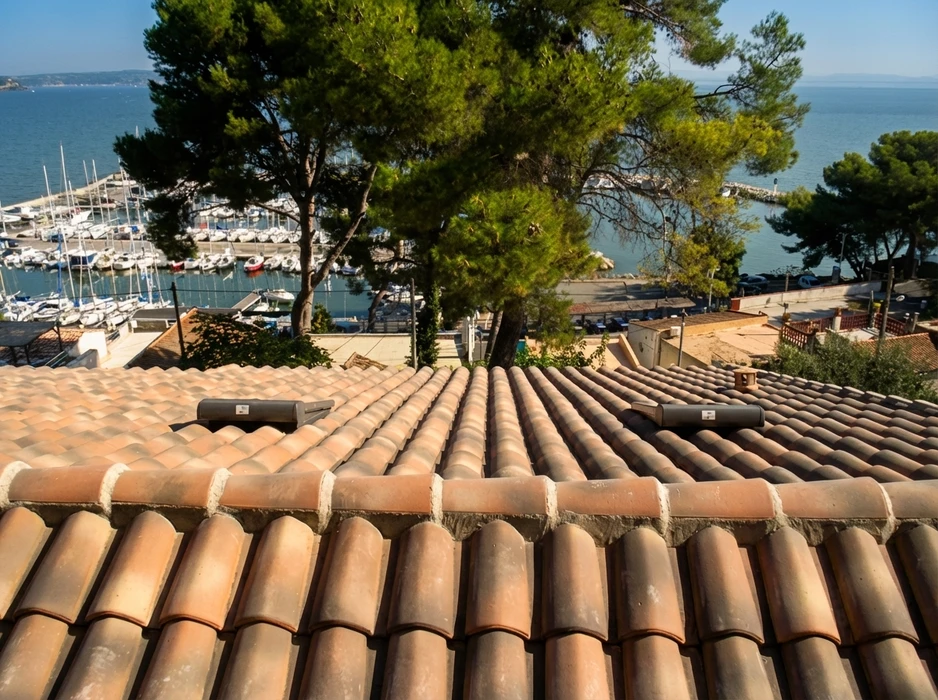 Ouvrier couvreur en tenue de protection travaillant sur toiture en tuiles mécaniques grises, ciel bleu dégagé